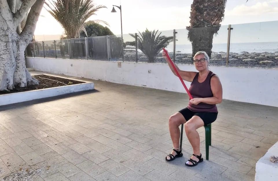 Person deltar i utomhusträning inom klimatvård på Teneriffa. Sittande övning med träningsband i varmt klimat som del av rehabilitering vid reumatisk sjukdom.