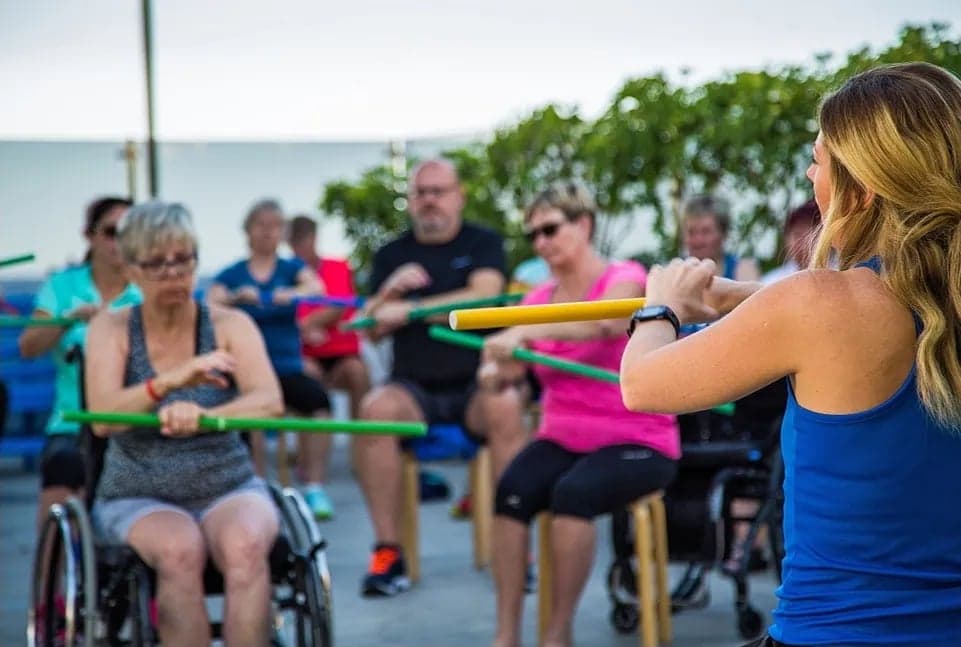 Gruppträning utomhus inom klimatvård. Personer med reumatisk sjukdom tränar sittande med stavar som del av multimodal rehabilitering i varmt klimat.